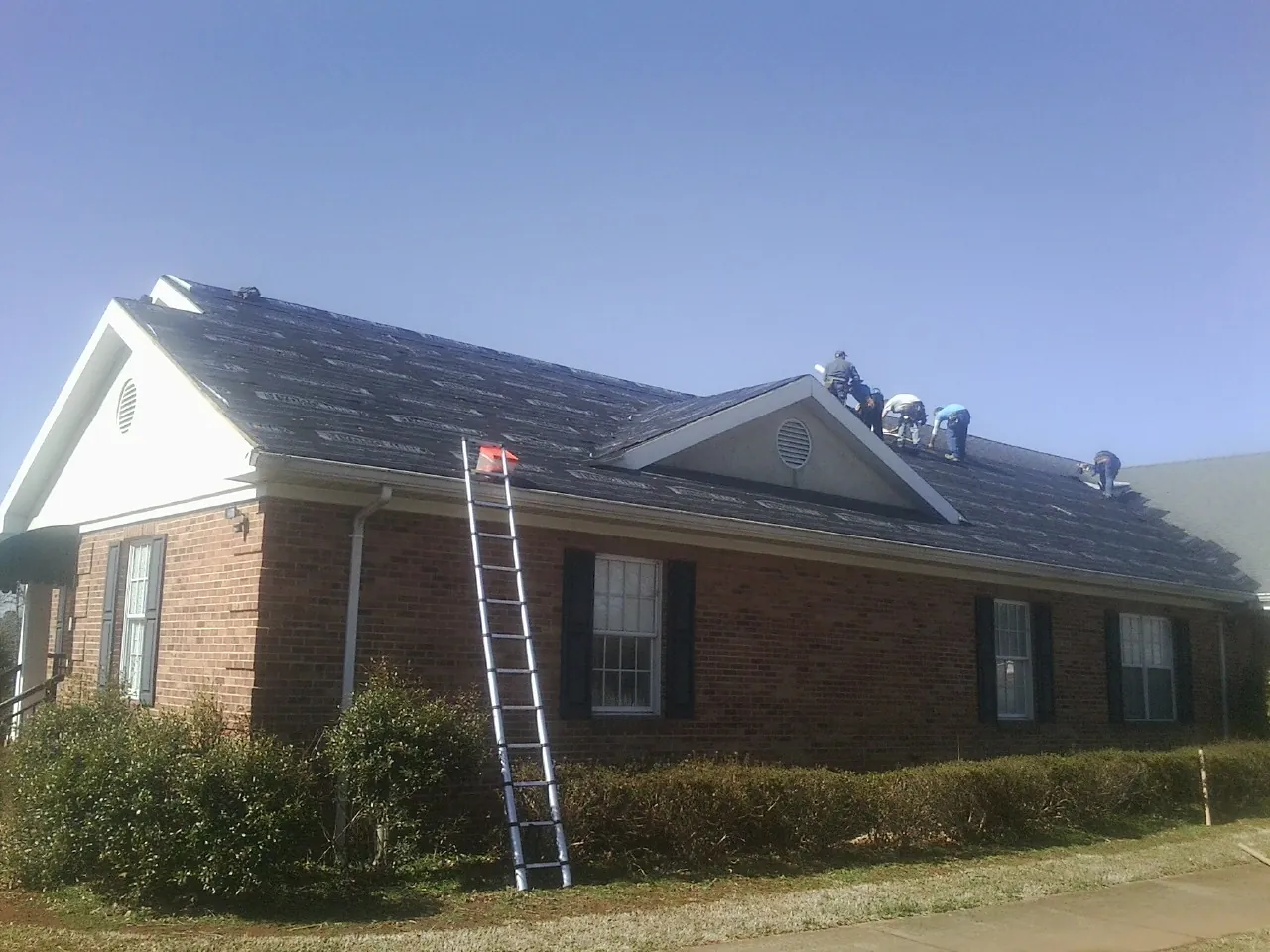Crew tearing shingles off a brick ranch roof with a ladder set — Emergency Roof Repair work by our crew in Manor