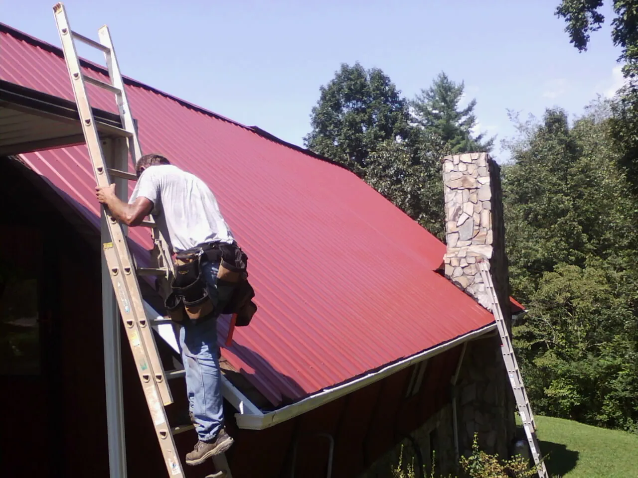 Roofer on a ladder beside a red metal roof and stone chimney — Soffit & Fascia Repair by Roofing Brothers in Manor